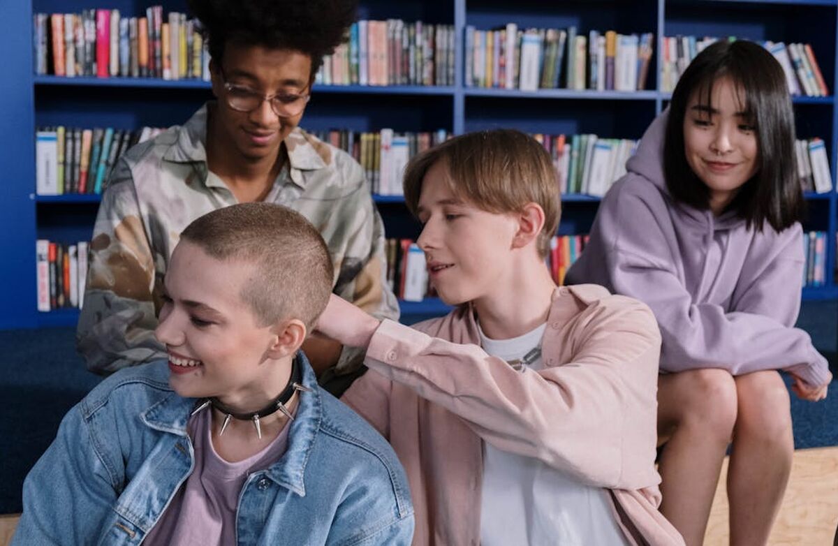 Bildet viser fire ungdommer som sitter sammen foran en bokhylle i det som ser ut som et bibliotek. De virker avslappede og smiler. Én person med kort, barbert hår sitter foran, mens en annen legger hånden vennlig på skulderen deres. De to andre sitter