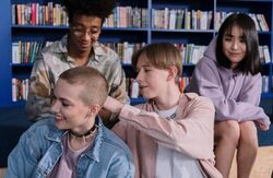 Bildet viser fire ungdommer som sitter sammen foran en bokhylle i det som ser ut som et bibliotek. De virker avslappede og smiler. Én person med kort, barbert hår sitter foran, mens en annen legger hånden vennlig på skulderen deres. De to andre sitter