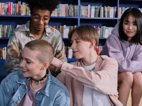 Bildet viser fire ungdommer som sitter sammen foran en bokhylle i det som ser ut som et bibliotek. De virker avslappede og smiler. Én person med kort, barbert hår sitter foran, mens en annen legger hånden vennlig på skulderen deres. De to andre sitter