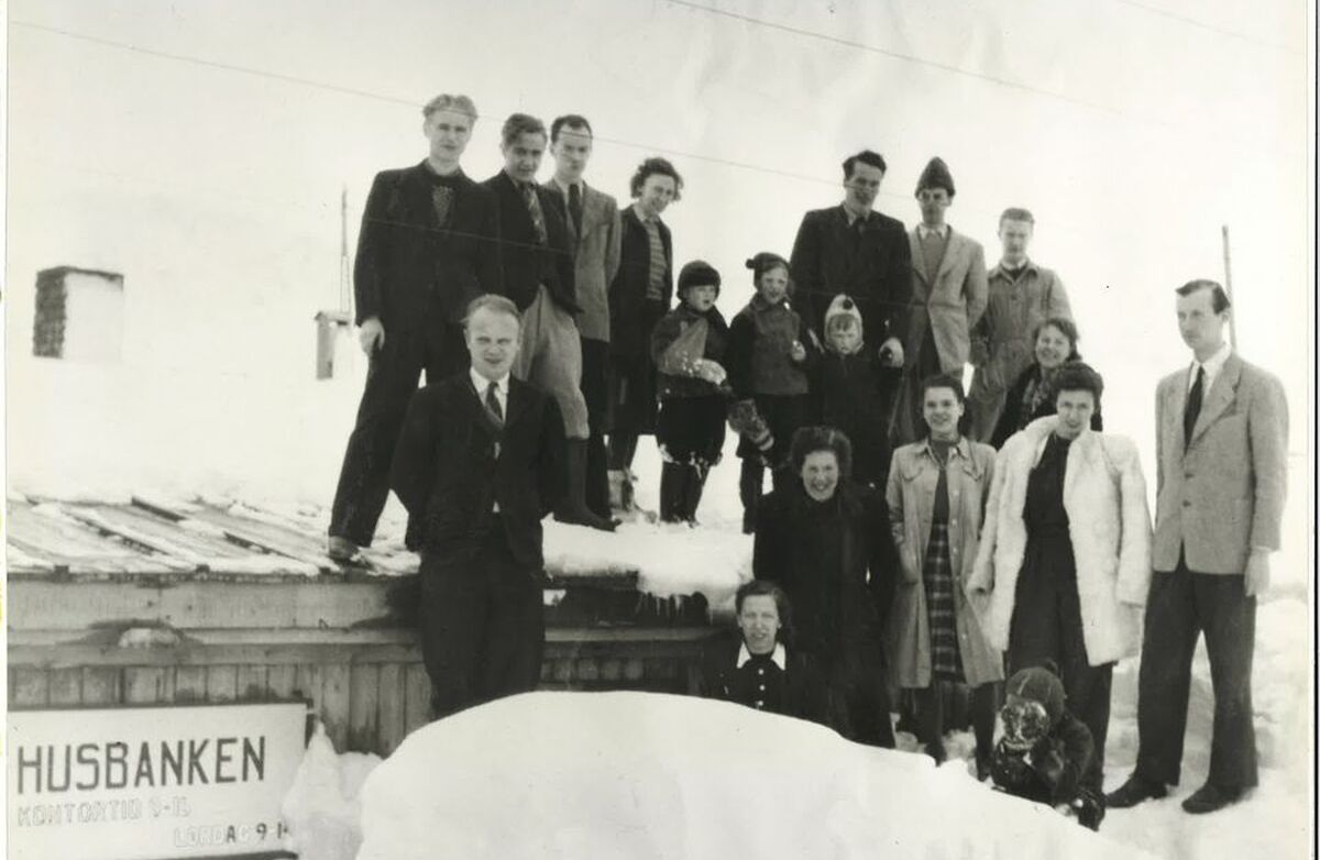 En gruppe på rundt femten personer står samlet utendørs i snøen foran en bygning. Flere står på taket. På veggen henger et skilt med teksten «Husbanken». Bildet er svart-hvitt og ser ut til å være fra midten av 1900-tallet.