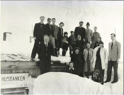 En gruppe på rundt femten personer står samlet utendørs i snøen foran en bygning. Flere står på taket. På veggen henger et skilt med teksten «Husbanken». Bildet er svart-hvitt og ser ut til å være fra midten av 1900-tallet.