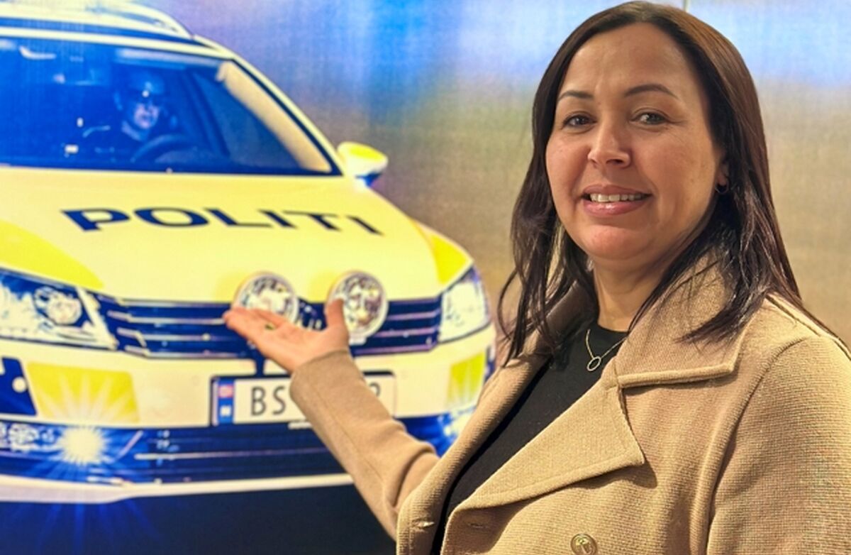 En kvinne står foran et bilde av en norsk politibil med blålys. Hun smiler og holder hånden frem som om hun presenterer bilen. På bilen står ordet «POLITI», og lysene gir en blå glød i bakgrunnen.