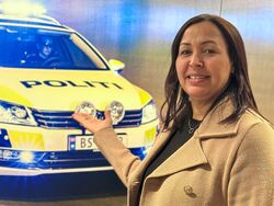 En kvinne står foran et bilde av en norsk politibil med blålys. Hun smiler og holder hånden frem som om hun presenterer bilen. På bilen står ordet «POLITI», og lysene gir en blå glød i bakgrunnen.