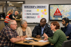 En mangfoldig gruppe voksne sitter i møte om beredskap, foran tavle med teksten “BEREDSKAP & SIKKERHET” og punkter om kriser, digital sikkerhet og førstehjelp, samt kartskjerm med rødt alarmsymbol.