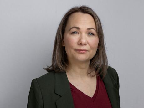 Kvinne med mørkt blondt hår, midtskill, dressjakke og burgunder V-genser ser alvorlig inn i kameraet.