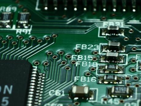 Bildet viser et nærbilde av et grønt kretskort med elektroniske komponenter, loddepunkter og tynne kobberbaner. Til venstre sees en del av en svart mikrochip, mens hvite merkinger og komponentkoder er trykt på kortet.