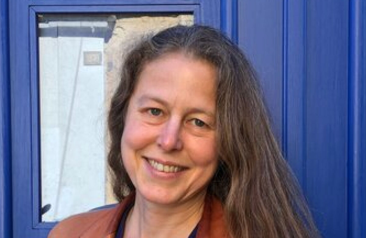 Kvinne med langt mellomblondt hår, smiler med hodet på skakke. En blå dør i bakgrunnen.