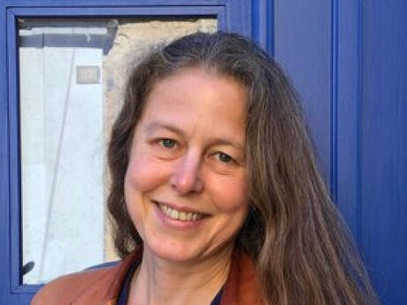 Kvinne med langt mellomblondt hår, smiler med hodet på skakke. En blå dør i bakgrunnen.