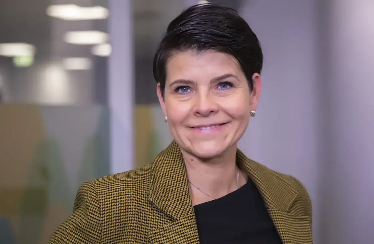 Kvinne med mørkt ganske kort hår og brune øyne. Brun jakke.