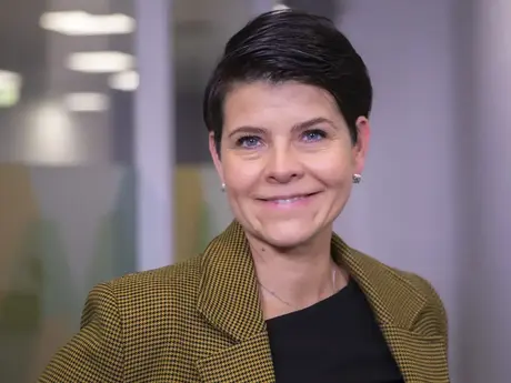 Kvinne med mørkt ganske kort hår og brune øyne. Brun jakke.