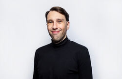 Ung mann med brunt hår og mørk genser smiler litt lurt. Nøytral bakgrunn.