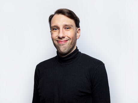 Ung mann med brunt hår og mørk genser smiler litt lurt. Nøytral bakgrunn.