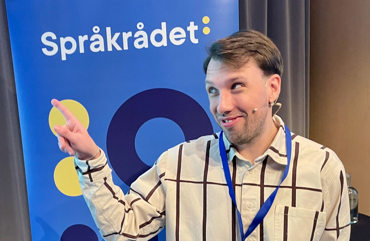 Ung mann med storrutete skjorte ser til siden og peker. I bakgrunnen ser vi en roll-up fra Språkrådet.