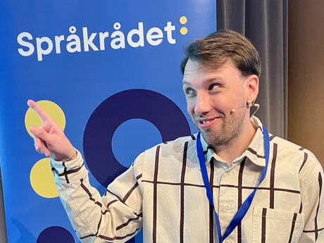 Ung mann med storrutete skjorte ser til siden og peker. I bakgrunnen ser vi en roll-up fra Språkrådet.