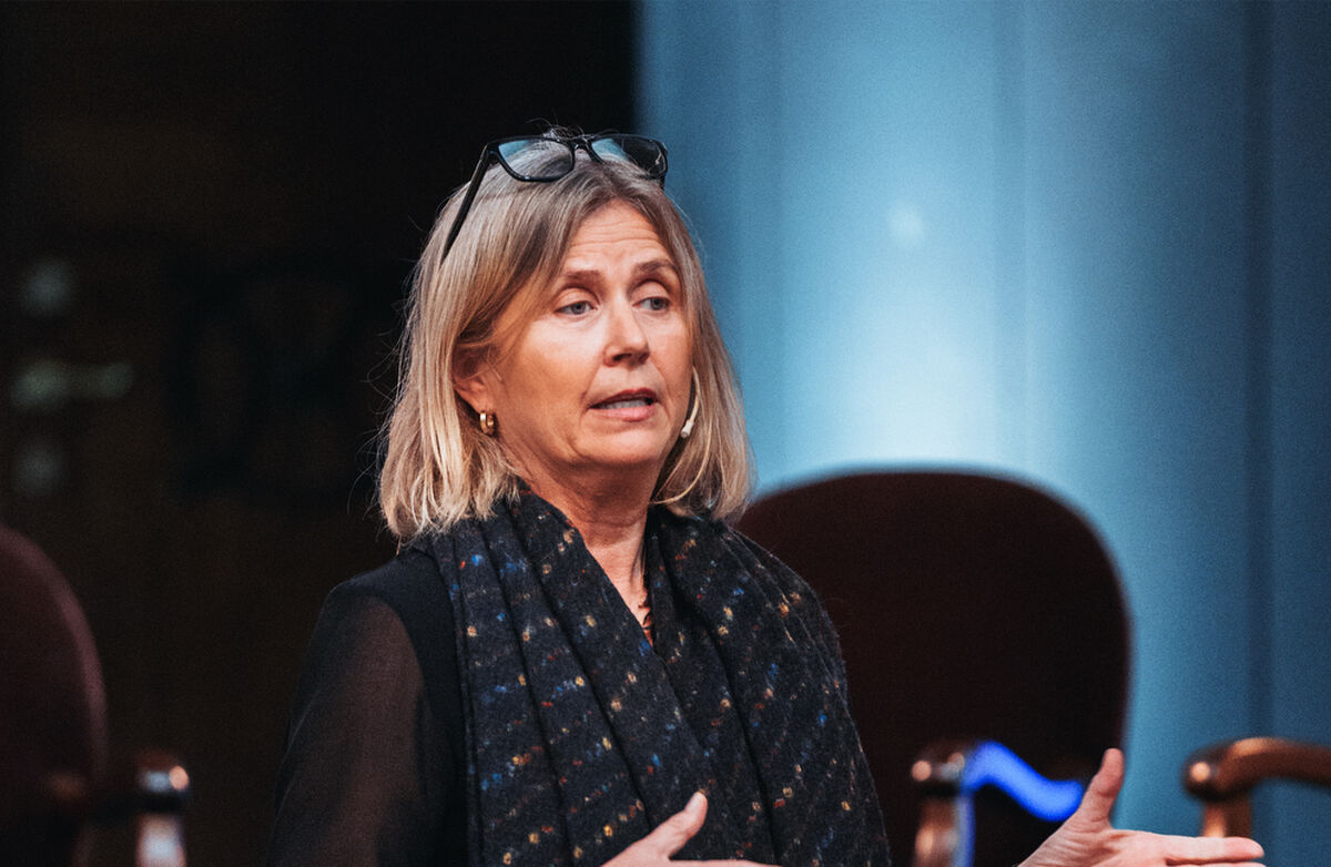 Middelaldrende kvinne med lyst halvlangt hår, midtskill, brillene er oppe på hodet. Hun ser til siden og holder hendene ut. Klærne er mørke. Hun står kanskje på en scene.