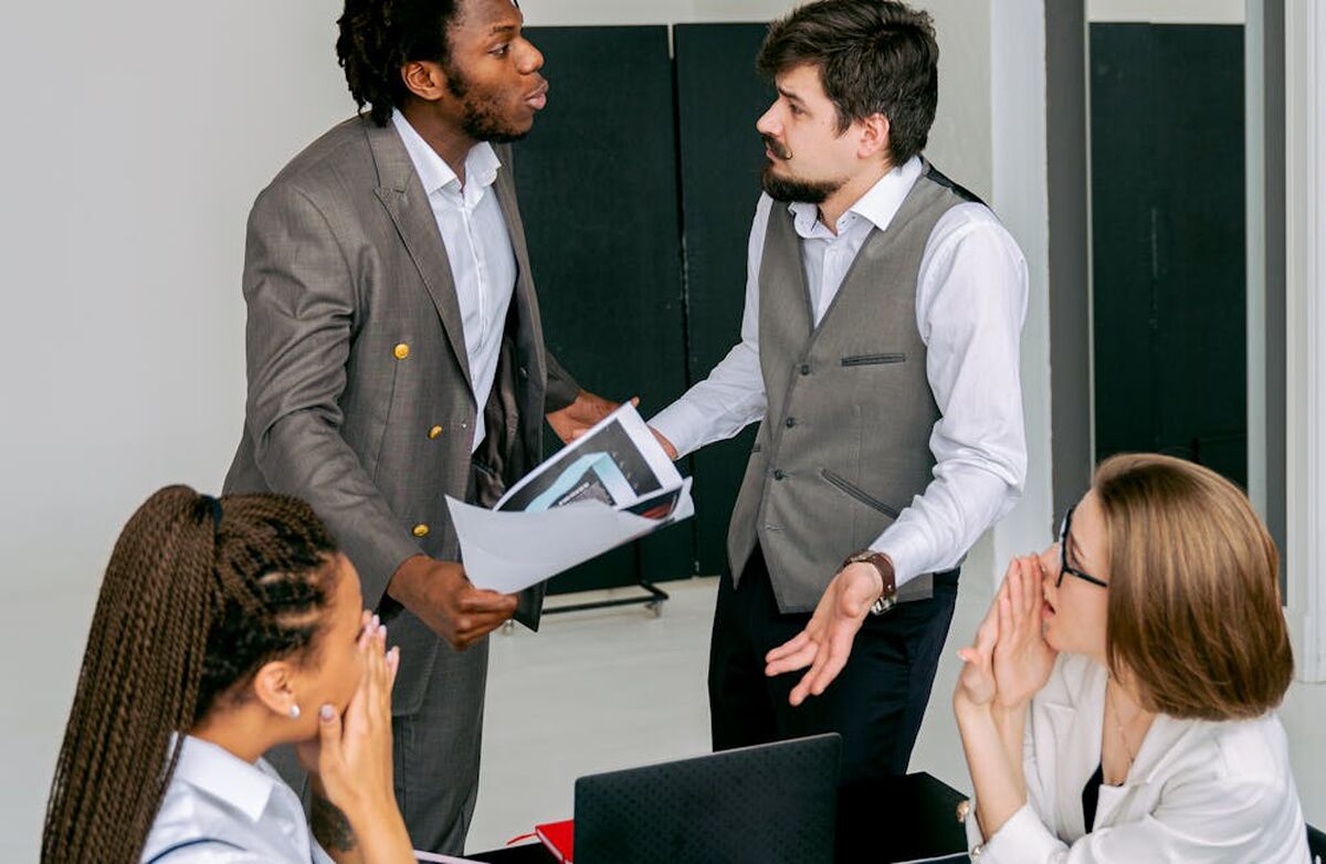 Fire kollegaer i et møte: To står og diskuterer tydelig og engasjert, mens to andre følger spent med rundt bordet. Bildet formidler konflikt, uenighet og krevende kommunikasjon på arbeidsplassen.