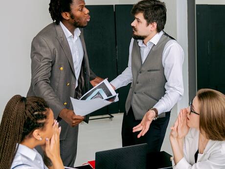 Fire kollegaer i et møte: To står og diskuterer tydelig og engasjert, mens to andre følger spent med rundt bordet. Bildet formidler konflikt, uenighet og krevende kommunikasjon på arbeidsplassen.