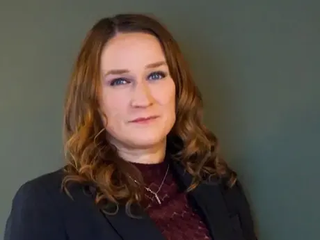 Kvinne med mørkt blondt hår med rødtone og blå øyne ser alvorlig inn i kameraet. Nøytrale mørke klær og bakgrunn.