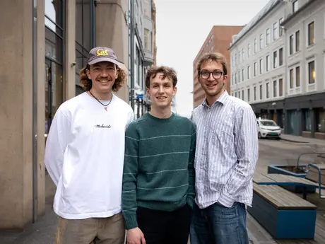 Bildet viser tre unge menn utendørs i et bymiljø. De smiler mot fotografen, og er uformelt kledd. En av dem har cap, en har briller.