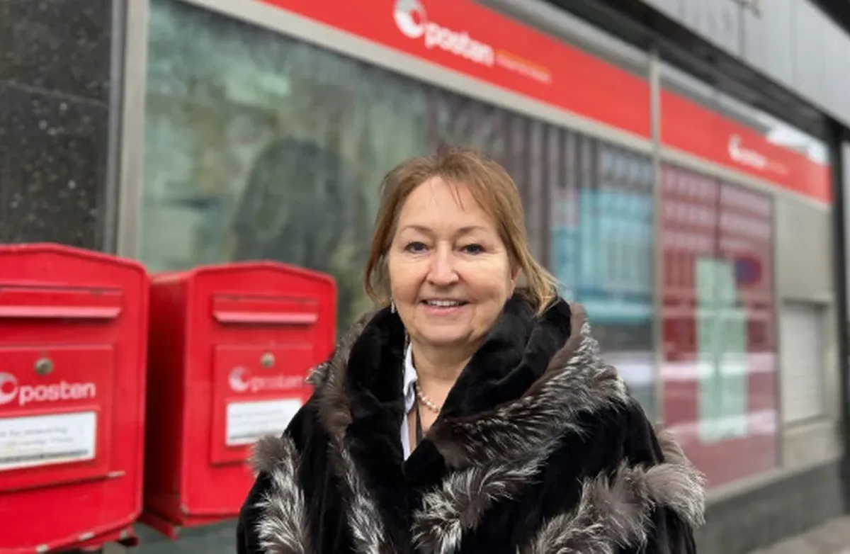 Gunn Marit Helgesen, styreleder i KS, mener at På dørterskelen bør innføres som en del av Postens omstilling. Foto: Turid Edvardsen En kvinne står smilende utenfor et postkontor eller Posten-lokale, ved siden av to røde postkasser. Hun har mørk kåpe med pelsdetaljer, og bygget i bakgrunnen er lett uskarpt.