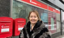En kvinne står smilende utenfor et postkontor eller Posten-lokale, ved siden av to røde postkasser. Hun har mørk kåpe med pelsdetaljer, og bygget i bakgrunnen er lett uskarpt.