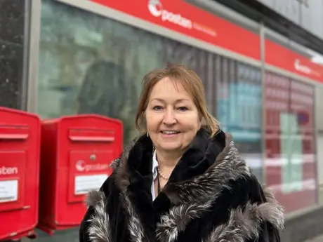 En kvinne står smilende utenfor et postkontor eller Posten-lokale, ved siden av to røde postkasser. Hun har mørk kåpe med pelsdetaljer, og bygget i bakgrunnen er lett uskarpt.