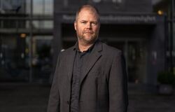 Middelaldrende mann med lite hår fotografert utendørs med en kontorbygning i bakgrunnen. Mørkt og litt dystert.