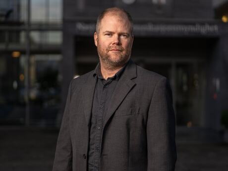 Middelaldrende mann med lite hår fotografert utendørs med en kontorbygning i bakgrunnen. Mørkt og litt dystert.