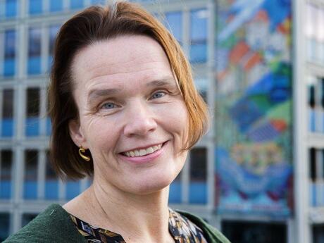 Middelaldrende kvinne smiler mot kameraet. Bildet er tatt utendørs i et bymiljø.