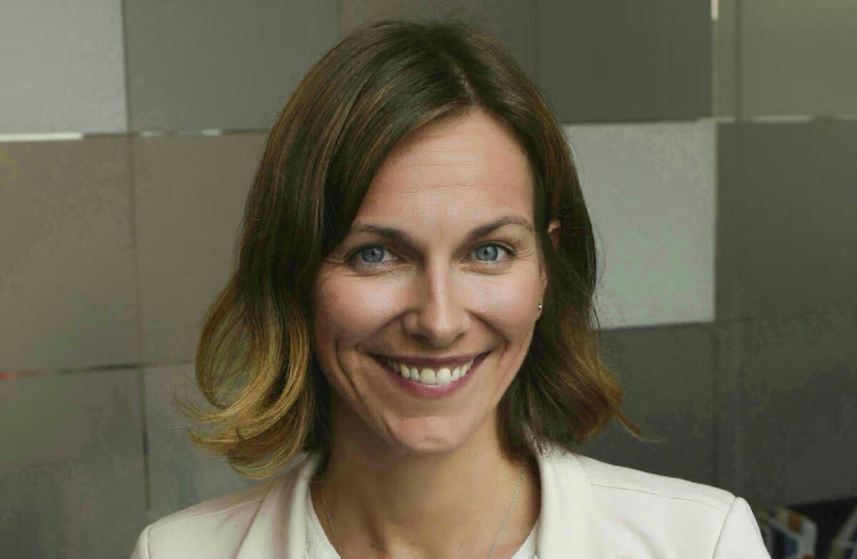 Kvinne med mellomblondt hår, hvit dressjakke og en hvit bluse/trøye innenfor. Hun smiler mot kamera.