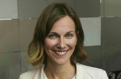 Kvinne med mellomblondt hår, hvit dressjakke og en hvit bluse/trøye innenfor. Hun smiler mot kamera.