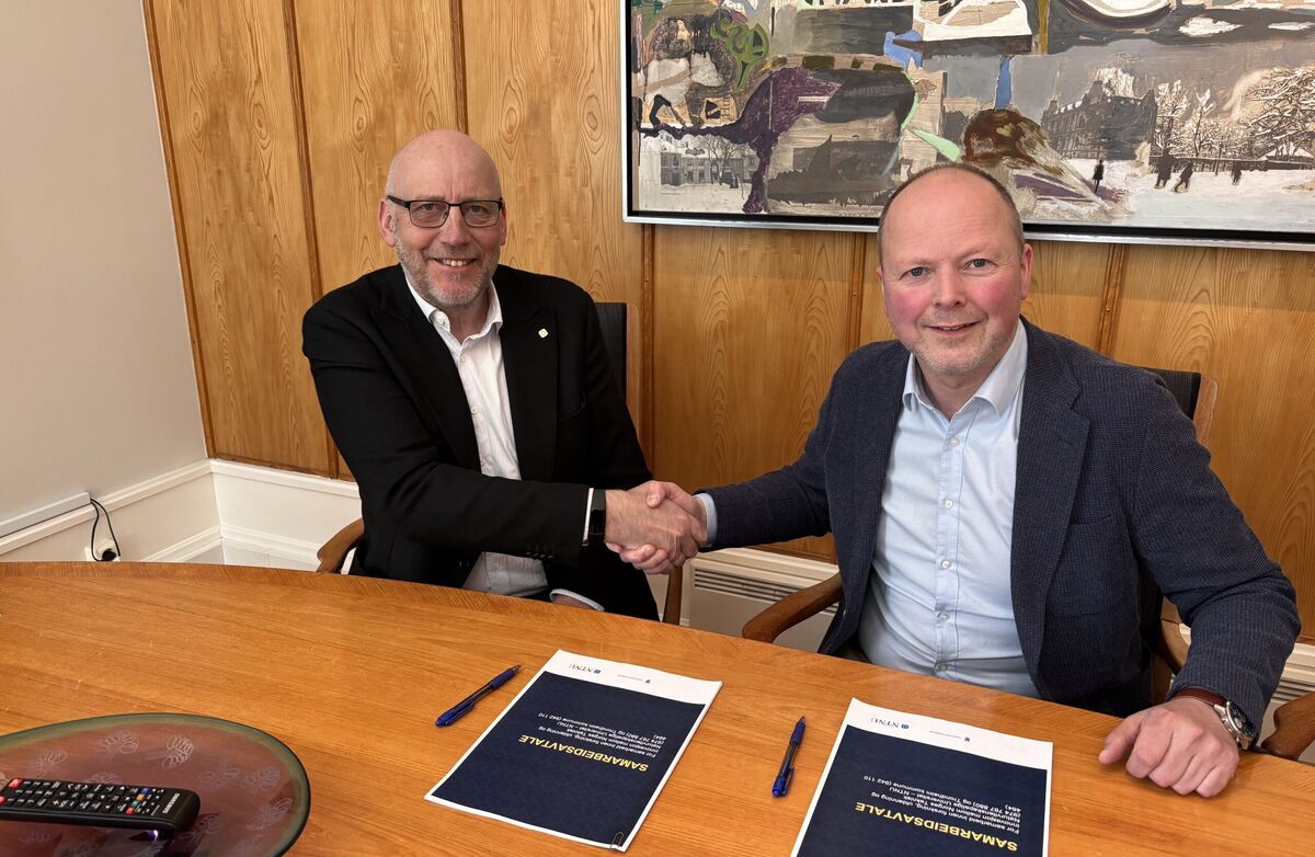 To middelaldrende menn i dress sitter bak et bord, gir et håndtrykk og ser inn i kameraet og smiler