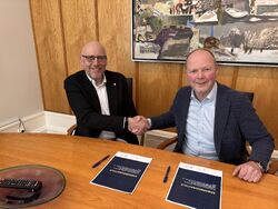 To middelaldrende menn i dress sitter bak et bord, gir et håndtrykk og ser inn i kameraet og smiler