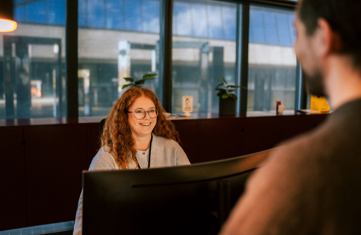 Kvinne med rødt langt hår smiler mot en person som står bak en skranke. Hun sitter ved en skjerm.