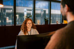 Kvinne med rødt langt hår smiler mot en person som står bak en skranke. Hun sitter ved en skjerm.