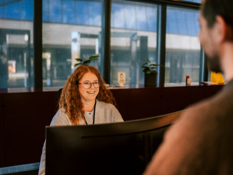 Kvinne med rødt langt hår smiler mot en person som står bak en skranke. Hun sitter ved en skjerm.