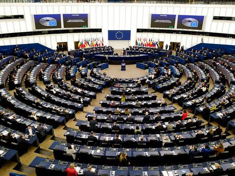 Bildet viser Europaparlamentets møtesal, som er rund. Vi ser fra høyden og ned mot scenen med diverse flagg, og medlemmene bakfra.