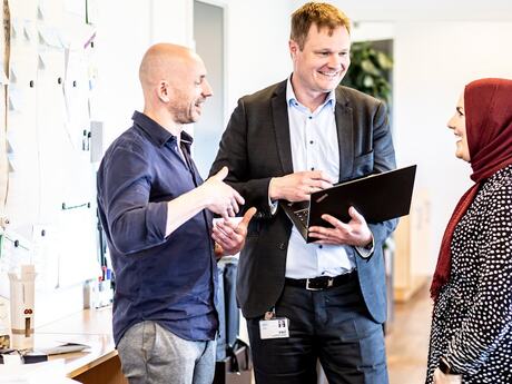 Tre kolleger står i et lyst kontormiljø og snakker sammen. En mann holder en laptop, en annen gestikulerer, og en kvinne i hijab smiler. På veggen bak henger lapper og notater.