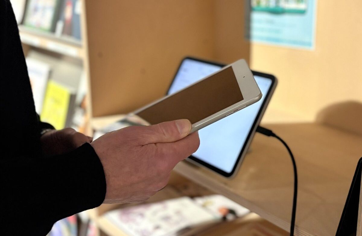 Hånd og deler av en arm. Holder en mobil. En skjerm i bakgrunnen.