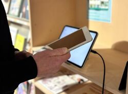 Hånd og deler av en arm. Holder en mobil. En skjerm i bakgrunnen.