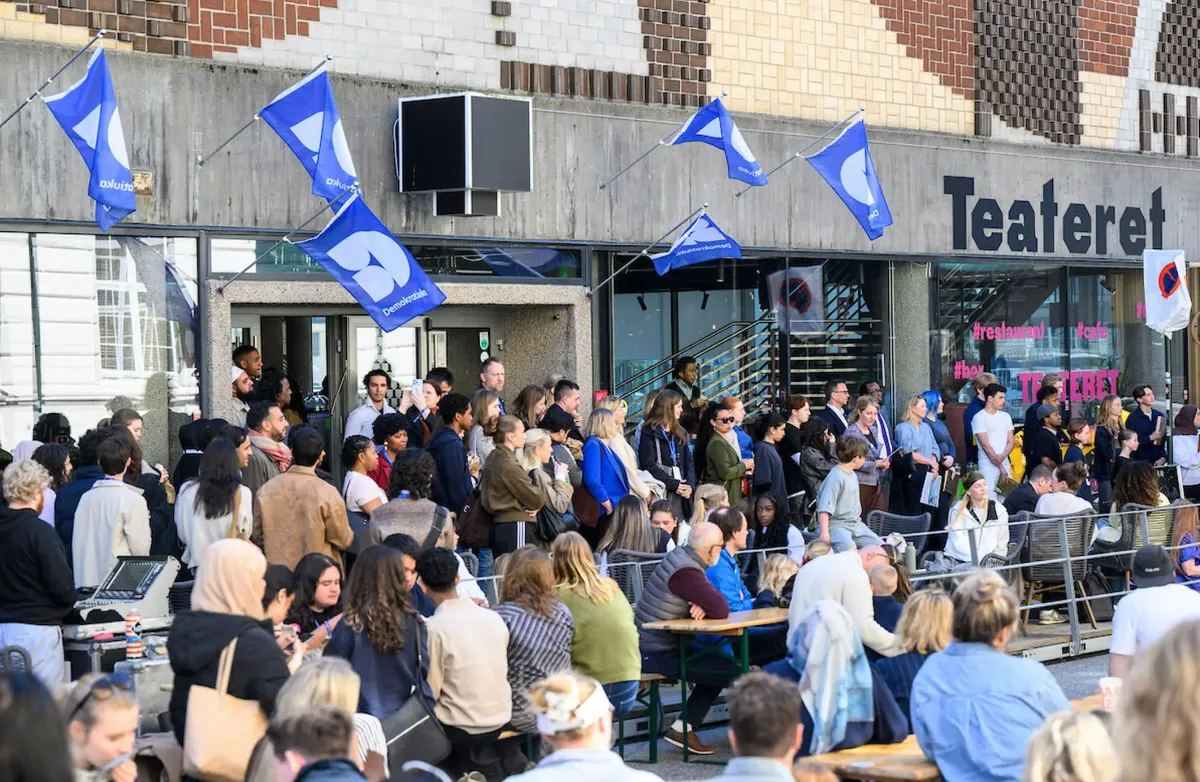Demokratiuka er en langsiktig satsing på å styrke hverdagsdemokratiet – i klasserom, i frivilligheten, i mediene og i møtet mellom innbyggere og folkevalgte, skriver artikkelforfatteren. Bildet er fra ett av arrangementene i år. Foto: Kjell Inge Søreide Mange mennesker samlet, noen står og noen sitter, det er blå vimpler festet på betongveggen med teksten Demokratiuka.
