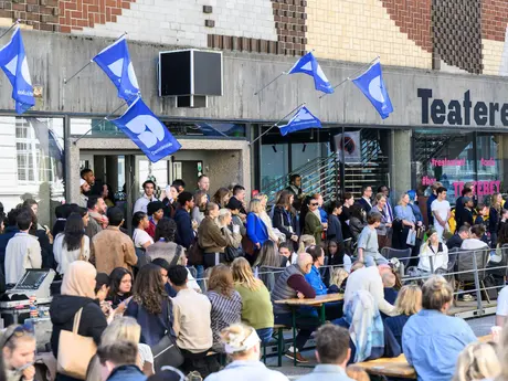 Mange mennesker samlet, noen står og noen sitter, det er blå vimpler festet på betongveggen med teksten Demokratiuka.