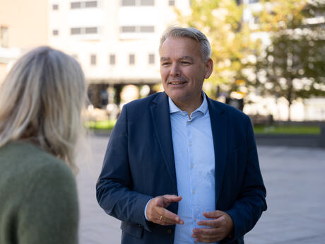 Bildet vis Jan Hjelle utendørs i samtale med en kvinne som vi ser bakfra.