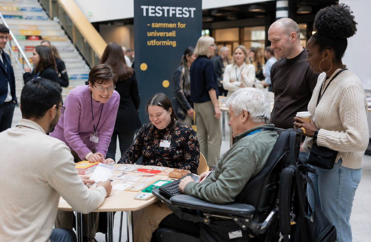 En mangfoldig gruppe mennesker møtes rundt et bord på Testfest for å teste og lære om universell utforming i et inkluderende miljø.