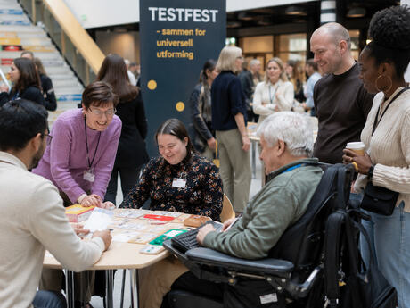 En mangfoldig gruppe mennesker møtes rundt et bord på Testfest for å teste og lære om universell utforming i et inkluderende miljø.