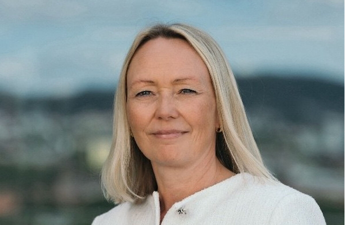 Kvinne med langt lyst hår, og en hvit jakke der vi bare ser deler av skuldrene. Hun myser litt mot kamera. Bakgrunnen er dus, men vi ser at hun er utendørs.