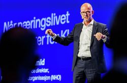 Skallet mann med dress og hvit skjorte på scenen. Armene litt utover.