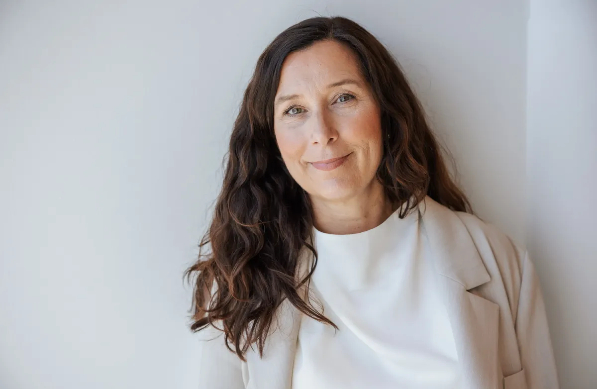– Dette er noe helt nytt og er foreløpig en betaversjon. Med denne assistenten kan du ha en dialog med læringsinnholdet og sparre med selve kildegrunnlaget, forklarer Christine Hassel Kristoffersen, leder for kompetanse i Digital Norway. Foto: Johnny Vaet Nordskog Portrett av en kvinne med langt, mørkt hår, kledd i lys blazer og hvit topp. Hun står mot en lys, nøytral bakgrunn og ser rolig mot kamera med et svakt smil.