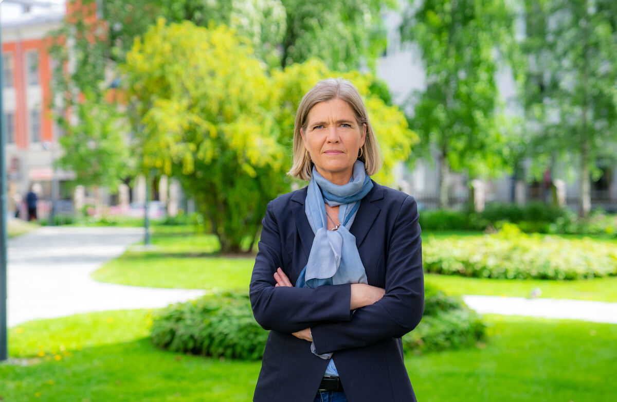 En alvorlig kvinne står ute i en grønn park med armene i kors. Hun har mørk blazer og lyst skjerf, og ser rett i kamera. Bak henne er trær, plen og bygninger i myk uskarphet.