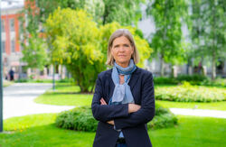 En alvorlig kvinne står ute i en grønn park med armene i kors. Hun har mørk blazer og lyst skjerf, og ser rett i kamera. Bak henne er trær, plen og bygninger i myk uskarphet.